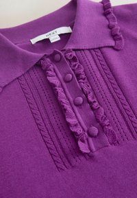 Haut en maille violet avec un col, des boutons recouverts de tissu, et des détails décoratifs en câble tricoté et volants le long de la patte de boutonnage.