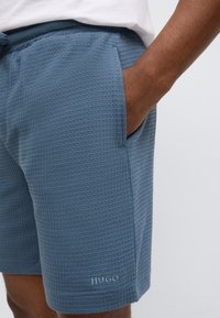 Blue textured shorts with a grid pattern, side pockets, and a logo detail on the hem. Elastic waistband for adjustable fit.