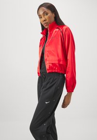 Sarkanais samta īsais žakets ar augstu apkakli un elastīgu jostasvietu, apvienots ar melniem jogger biksēm, kuru sānos ir pogas un Nike logo.