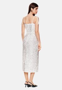 Weißes Midi-Kleid mit Allover-Pailletten, verstellbaren Spaghettiträgern, tailliertem Oberteil und seitlichem Schlitz. Kombiniert mit schwarzen Slingback-Schuhen.