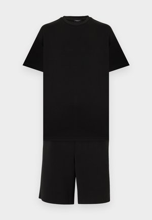 Fekete, rövid ujjú, kerek nyakú póló fekete, térdig érő rövidnadrággal, világosszürke egyszínű háttéren.