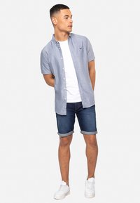 Blauwe chambray overhemd met korte mouwen en knoopsluiting, een witte t-shirt met ronde hals eronder, gecombineerd met opgerolde denim shorts en witte sneakers.