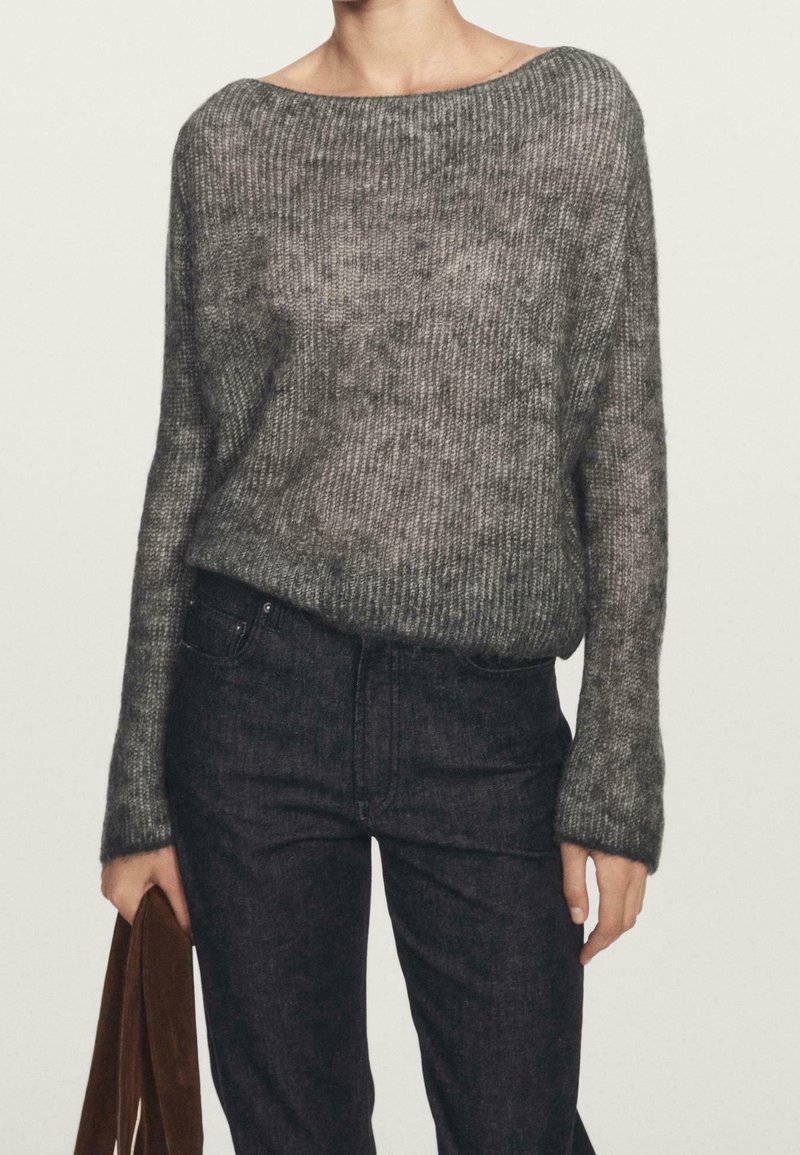 Personne portant un pull en maille gris ample et un jean taille haute foncé, tenant un sac marron à ses côtés.