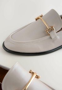 Lichtgrijze leren loafers met een ronde neus, voorzien van een gouden metalen accent en een zwarte zool. Gladde textuur met zichtbare stikseldetails.