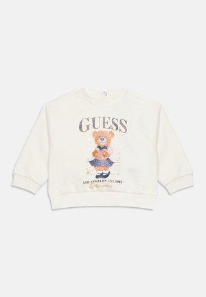 Hvid langærmet sweatshirt med en bamse iført nederdel og holdende en bog, mærket "GUESS Los Angeles Est. 1981 California."