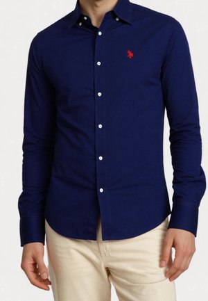 Mężczyzna ubrany w dopasowaną granatową, długorękawową koszulę na guziki z czerwonym haftowanym logo oraz beżowe spodnie, stojący z rękami swobodnie opuszczonymi.