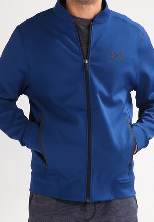 Mężczyzna noszący niebieską kurtkę na zamek błyskawiczny z czarnym logo Under Armour, ręce w kieszeniach i ciemne spodnie w wzory.