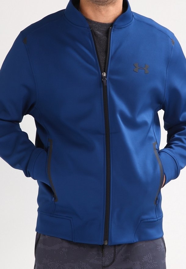 Homme portant une veste bleue zippée avec logo Under Armour noir, mains dans les poches et pantalon foncé à motifs.