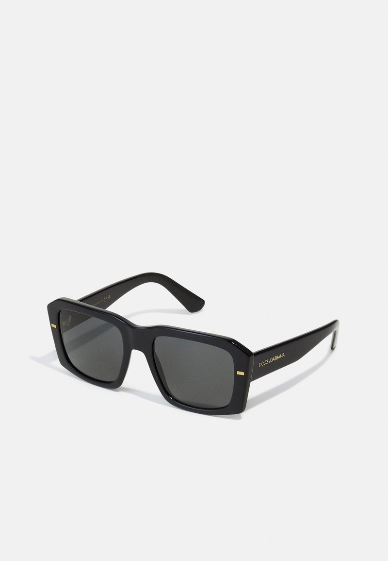 Ochelari de soare din plastic negru, cu lentile de culoare gri închis, de formă pătrată și accente aurii pe ramă. Numele mărcii este afișat pe lateral.