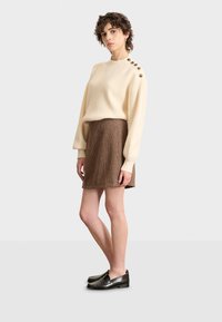 Crème ribbeltrui met knoopaccenten op de schouder, gecombineerd met een bruine gestructureerde A-lijn rok en zwarte instappers. Netjes, casual outfit.