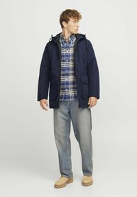 Temno modra kapuca parka z dvema sprednjima žepoma, nosi se čez karo srajco v modri in beli barvi, v kompletu z svetlim jeansom in bež čevlji.