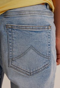 Světle modré denimové džíny mají design zadní kapsy ve tvaru jha s kontrastním prošíváním. Tkanina má měkkou, mírně opotřebovanou strukturu.