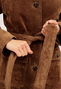 Brun ruskind frakke med bælte i taljen, der har store forlommer og mørke knapper. Teksturen er blød med en glat overflade.