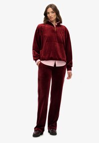 Sudadera de terciopelo burdeos con media cremallera y bolsillo frontal, combinada con pantalones a juego. El conjunto incluye una camisa rosa de contraste debajo.