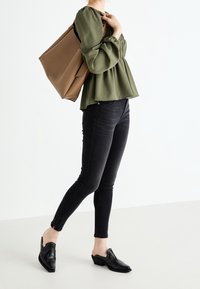 Haut péplum vert olive avec poignets élastiques, jean skinny noir et chaussures noires slip-on avec détails de design. Sac fourre-tout marron sur l'épaule.