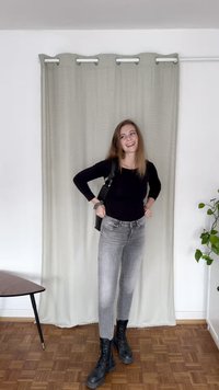 Top preto de mangas longas, jeans ajustados cinzentos, botas de combate pretas e uma bolsa de ombro preta; de pé contra uma cortina clara e um chão de madeira.