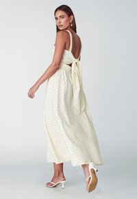 Vestido midi color crema con lunares negros, espalda descubierta, lazos ajustables en la cintura y falda acampanada. Zapatos de tacón alto blancos visibles.