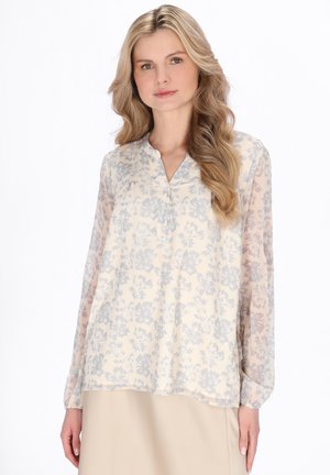 Kobieta o długich blond włosach, ubrana w jasną, kwiecistą bluzkę z przezroczystymi rękawami i beżową spódnicę, stoi na białym tle.