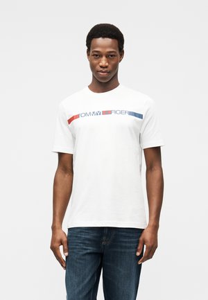 Νέος άνδρας στέκεται φορώντας ένα λευκό t-shirt Tommy Hilfiger με κόκκινες και μπλε ρίγες και σκουρόχρωμο μπλε τζιν, μπροστά σε απλό ανοιχτό φόντο.