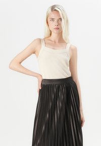 Débardeur en maille beige avec des bretelles larges et un décolleté carré, assorti à une jupe plissée noire avec une texture lisse et brillante.