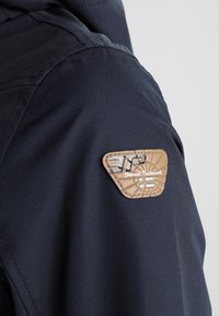 Manche de veste bleu foncé avec un patch triangulaire beige portant le texte « Finnish Trademark » et un motif embossé subtil.