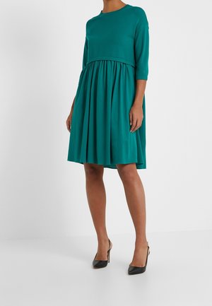 Femme portant une robe bleu sarcelle mi-longue avec manches trois-quarts et jupe plissée, associée à des chaussures slingback noires à talons hauts.