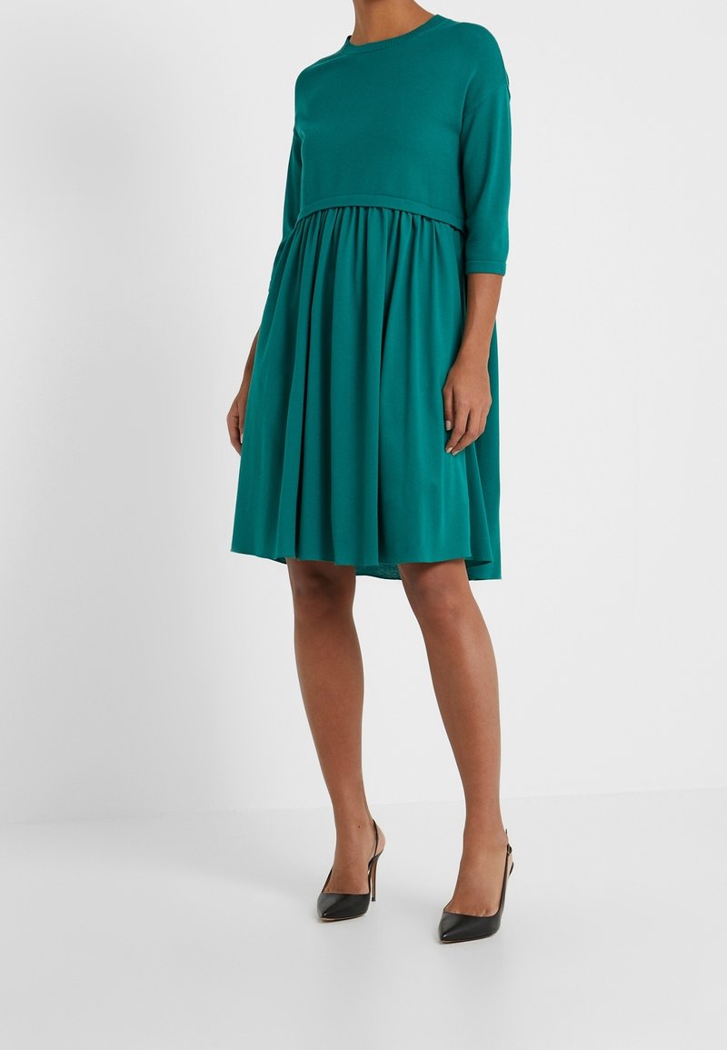 Femme portant une robe bleu sarcelle mi-longue avec manches trois-quarts et jupe plissée, associée à des chaussures slingback noires à talons hauts.