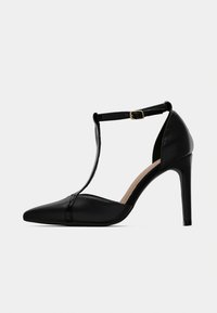 Μαύρο γυναικείο παπούτσι με τακούνι, T-strap με μυτερή μύτη και χρυσή αγκράφα στο λουράκι του αστραγάλου, απεικονιζόμενο σε πλαϊνή όψη σε γκρι φόντο.