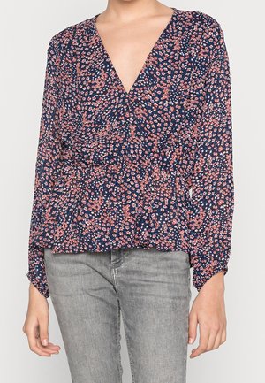 Kvinde iført en marineblå slå-om-bluse med lange ærmer med pink og hvid blomsterprint, kombineret med lysegrå jeans.