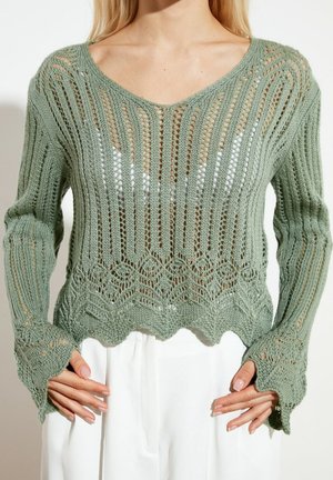 Kvinde iført en lysegrøn, åbent strikket sweater med bølgekant og lange ærmer over en hvid top, kombineret med hvide bukser.