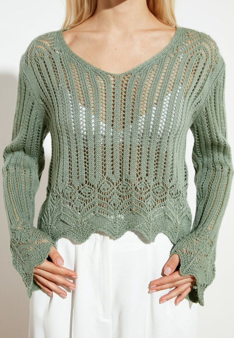 Femme portant un pull en maille légère vert clair à bords festonnés et manches longues, par-dessus un haut blanc, associé à un pantalon blanc.