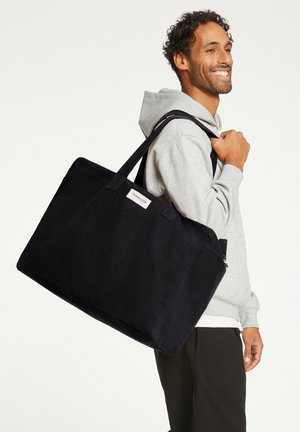 Black fabric tote bag with sturdy handles, featuring a rectangular shape and a front logo patch. Textured surface with a matte finish.