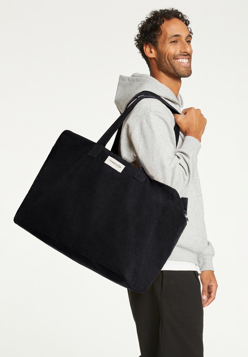 Sac en toile noir avec de solides poignées, de forme rectangulaire et orné d'un patch logo sur l'avant. Surface texturée avec un fini mat.