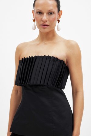 Woman wearing strapless black pleated top and large amber and white drop earrings against plain white background.