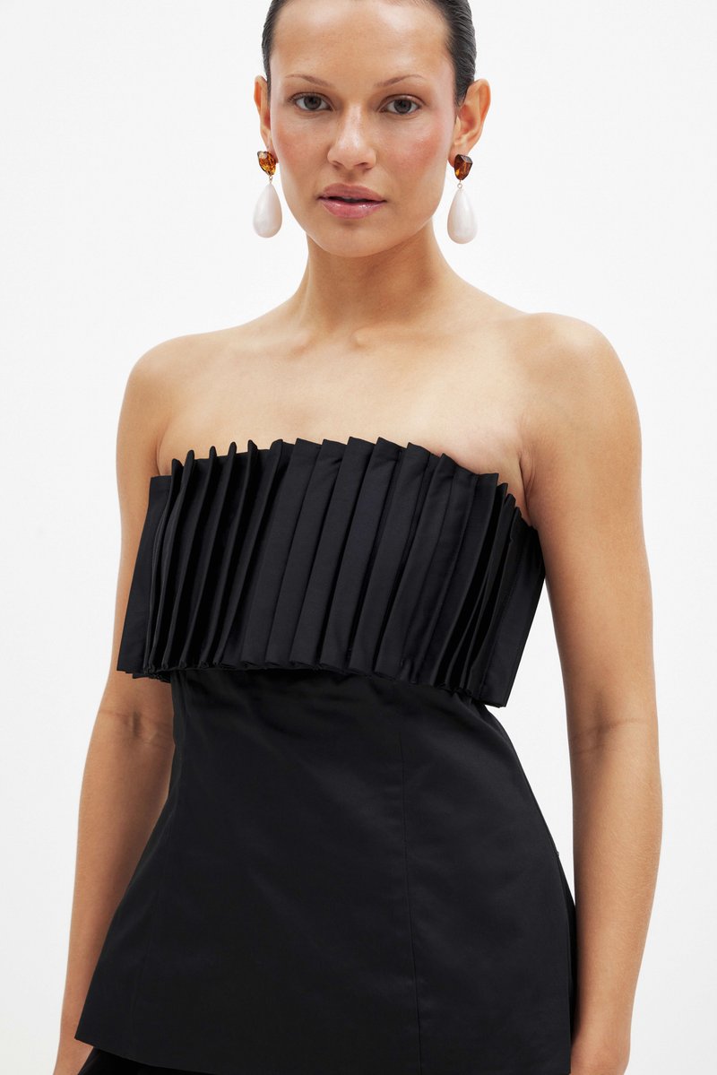 Kobieta nosząca bezramną, czarną plisowaną górę oraz duże kolczyki w kształcie kropli z bursztynu i białego koloru na tle jednolitego białego tła.