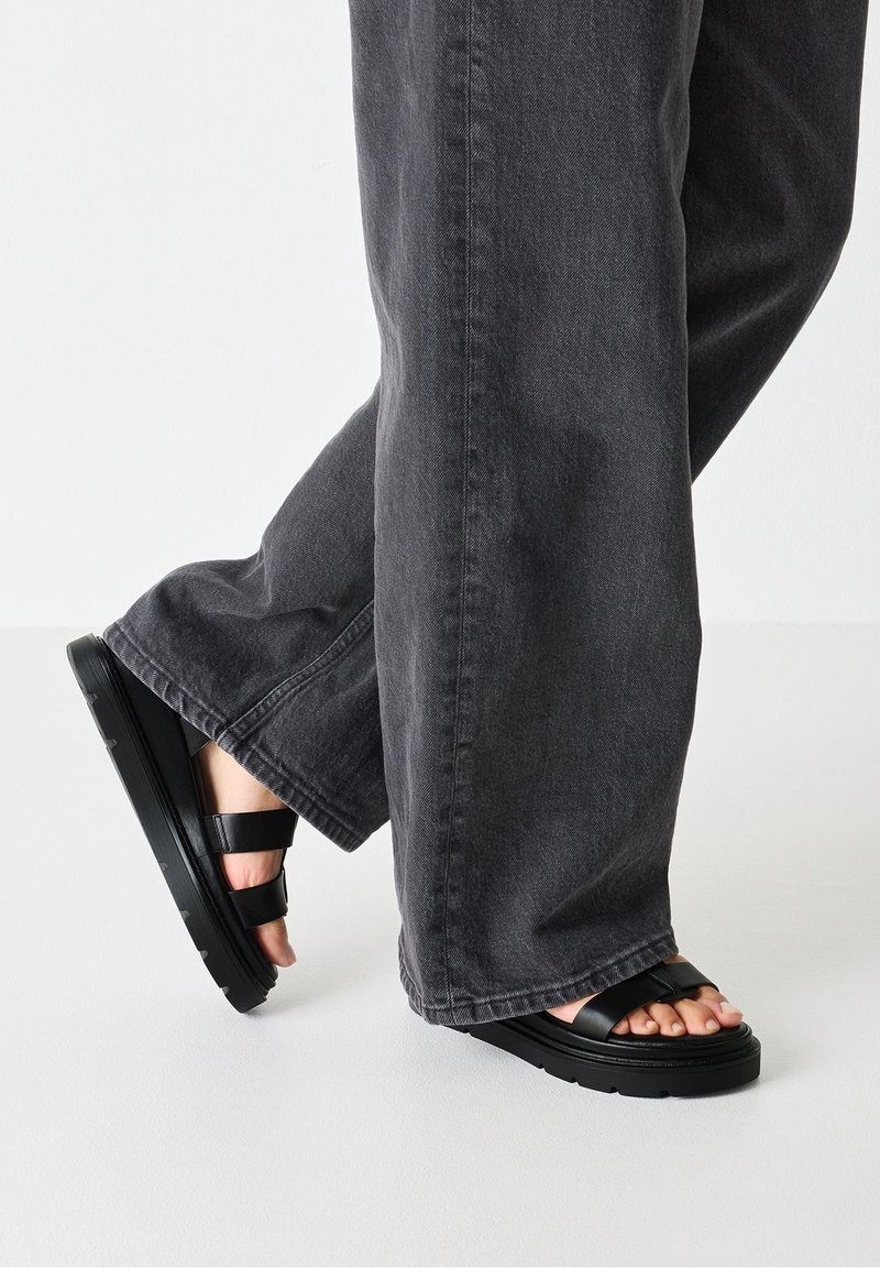 Pieds portant des sandales noires à semelles épaisses et à brides, associés à un pantalon large et sombre sur un fond clair uni.