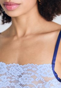 Brassière en dentelle lavande avec des bords festonnés et des bretelles satinées bleues, présentant des motifs floraux et une texture transparente.