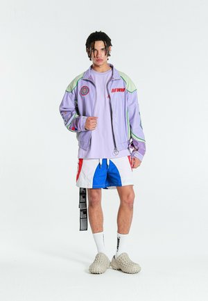 Joven con el cabello trenzado que lleva una chaqueta lavanda, camisa a juego, pantalones cortos bicolor, calcetines blancos y zapatos beige sin cordones, de pie sobre un fondo blanco.