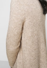 Beige stickad tröja med mjuk textur, med rund halsringning och långa ärmar, visad från sidan.