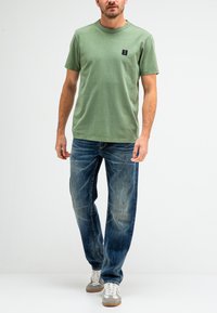 Groene katoen T-shirt met een ronde hals en een klein zwart logo, gecombineerd met blauw versleten denim jeans en grijze sneakers.
