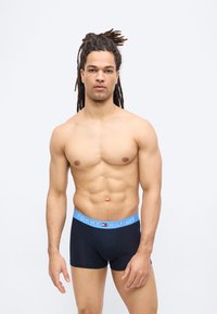 Μυώδης άνδρας με ντραντλοκς που φοράει μαύρα ανδρικά εσώρουχα με μπλε ζώνη Tommy Hilfiger, στέκεται μπροστά σε λευκό φόντο.