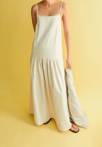 Vaalean beige maksi-mekko ohuilla olkaimilla, teksturoitua kangasta, kerrostettu hame ja mustat varvassandaalit, mekko yhdistetty vaaleaan takkiin.