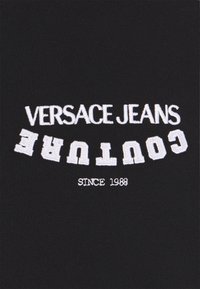 Černá tkanina s bílým vyšívaným textem "VERSACE JEANS COUTURE" a "SINCE 1988", přičemž název značky je obráceně vzhůru nohama.