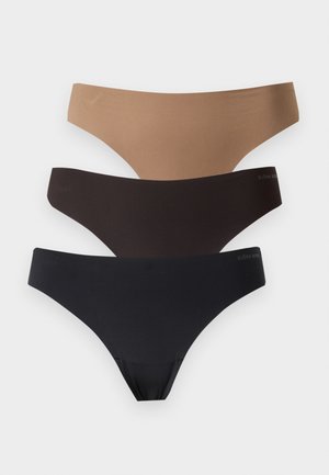 Jeune femme debout pieds nus portant un soutien-gorge noir et une culotte noire assortie sur un fond blanc uni.