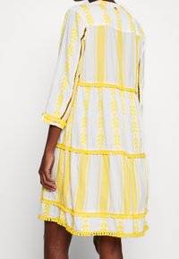 Weißes Kleid mit gelben Streifen und Blattmustern, mit Dreiviertelärmeln, einem gestuften Design und kontrastierenden gelben Abschlüssen.