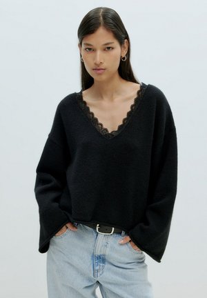 Schwarzer gestrickter Pullover mit V-Ausschnitt und Spitzenbesatz. Lockere Passform mit überschnittenen Schultern, kombiniert mit hellblauen Denim-Jeans und einem schwarzen Gürtel.