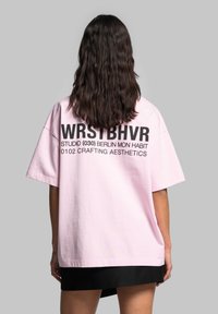 Růžové oversized tričko s černým textem na zádech, obsahující "WRSTBHV" a další detaily. Měkká bavlněná tkanina s uvolněným střihem.