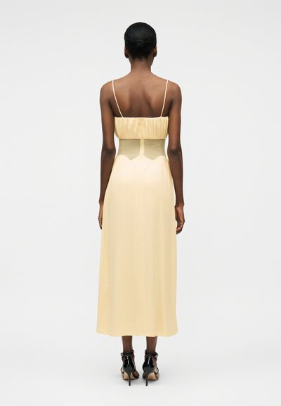 Γυναίκα με σκουρόχρωμο δέρμα και μαλλιά σε κότσο που φοράει ανοιχτοκίτρινο μίντι φόρεμα χωρίς μανίκια με λεπτές τιράντες και μαύρα ψηλοτάκουνα σανδάλια, στέκεται γυρισμένη προς τα πίσω.