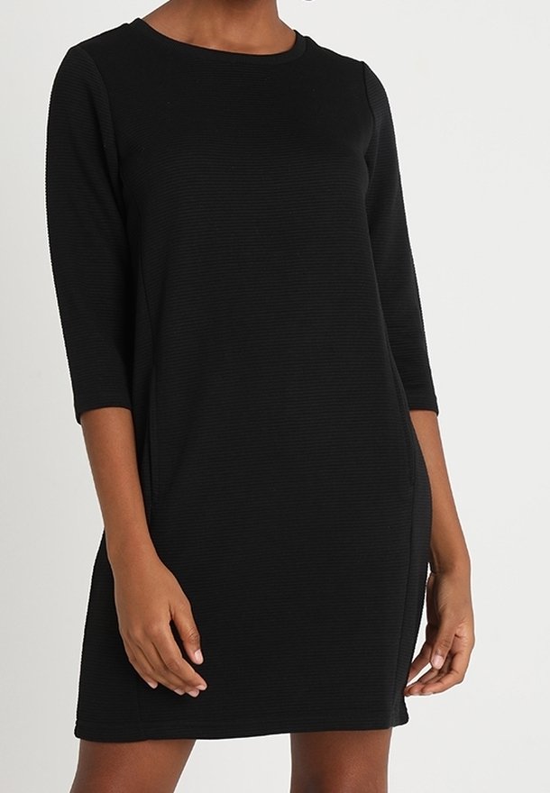 Femme portant une robe noire ajustée, longueur genoux, avec des manches trois-quarts et un col rond, sur un fond uni.