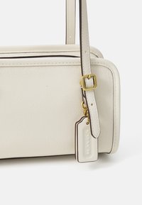 Balta oda crossbody kuprinė su tekstūruotu paviršiumi, rudos spalvos akcentais, auksinėmis detalėmis ir nuimamu žymekliu. Yra reguliuojamas diržas.